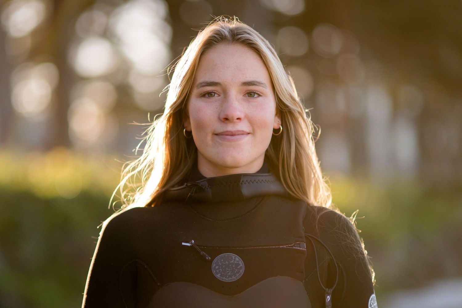 A surfer in a black wetsuit smiles warmly in golden sunlight with bokeh effect in the background.