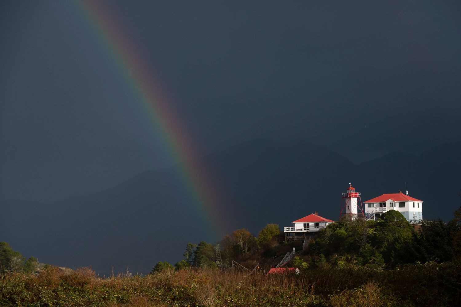 A lighthouse stands atop a hill against a dark stormy sky with a vibrant rainbow arcing overhead.