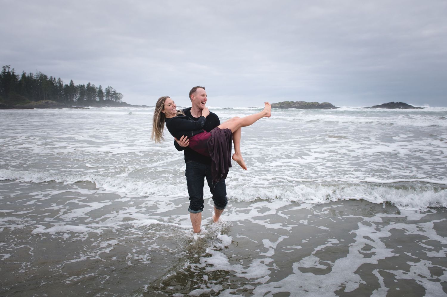 A couple shares a romantic moment on a stormy Pacific Northwest beach with crashing waves and cloudy skies.