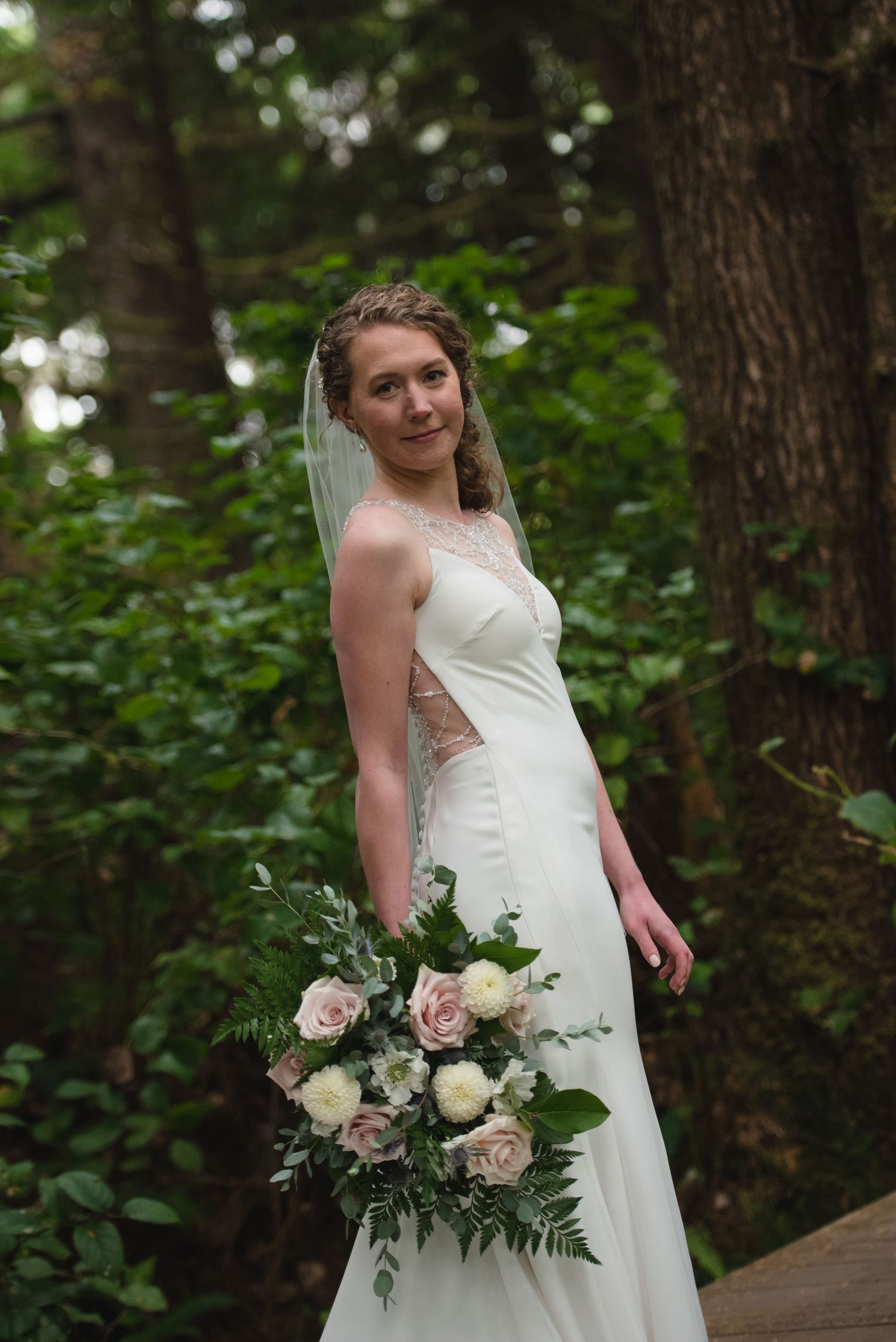 A bride in a white wedding dress holds a beautiful bouquet of pink and white roses in a wooded setting.