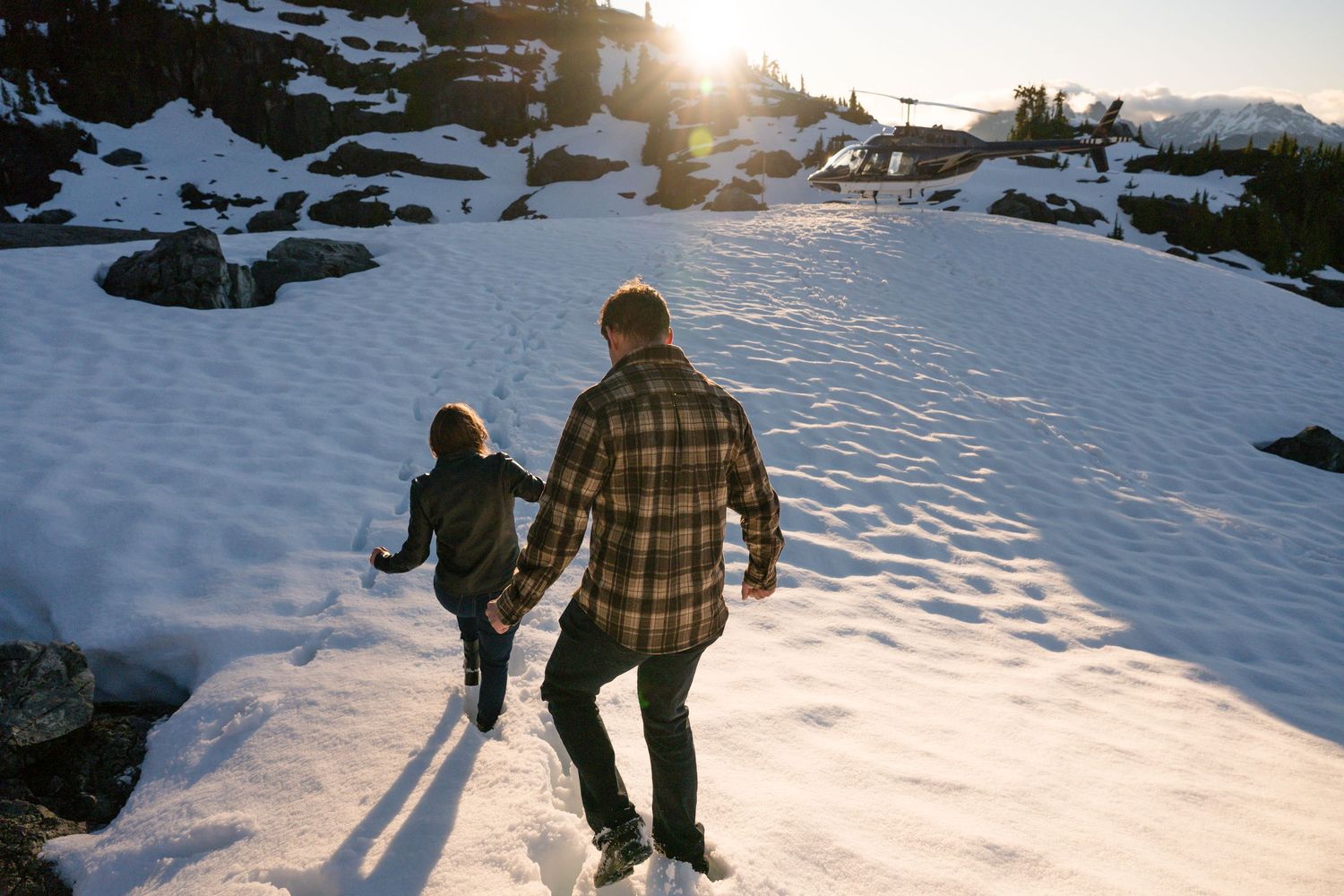 Couple holding hands walking up a snowy mountain trail toward a beautiful golden sunset.
