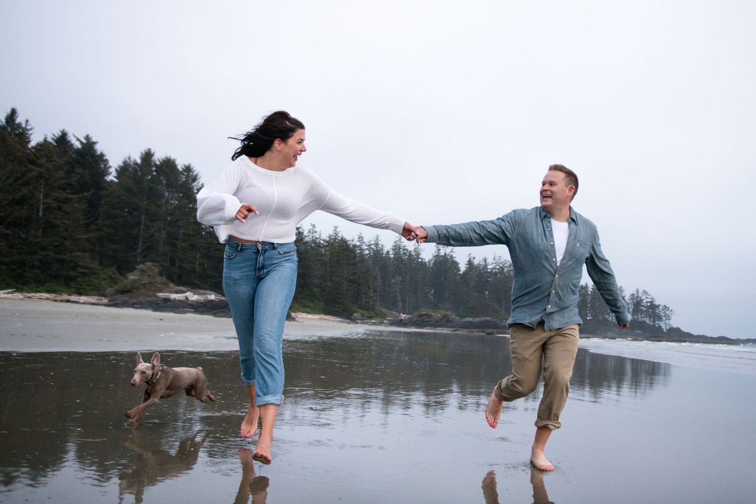 Two people laugh and play together during a beach photoshoot near a forested coastline.