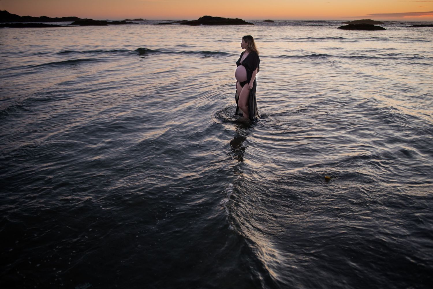 Silhouette standing in ocean waves during golden hour with dramatic sky.