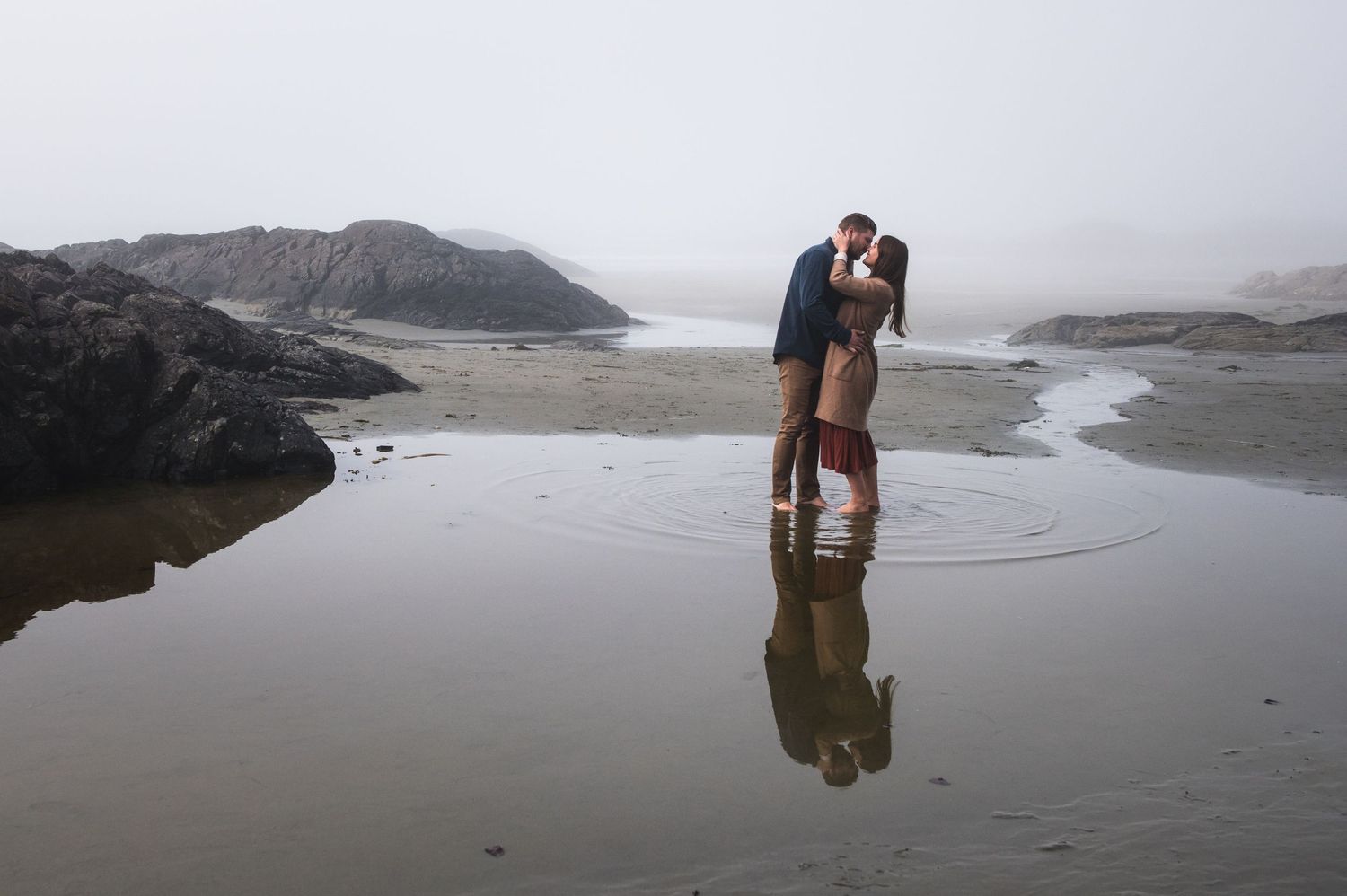 Figures walking near tide pools on remote beach with cliffs and fog.