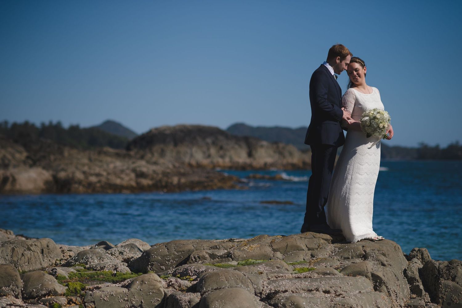 A couple shares an intimate moment on coastal rocks overlooking blue ocean waters.