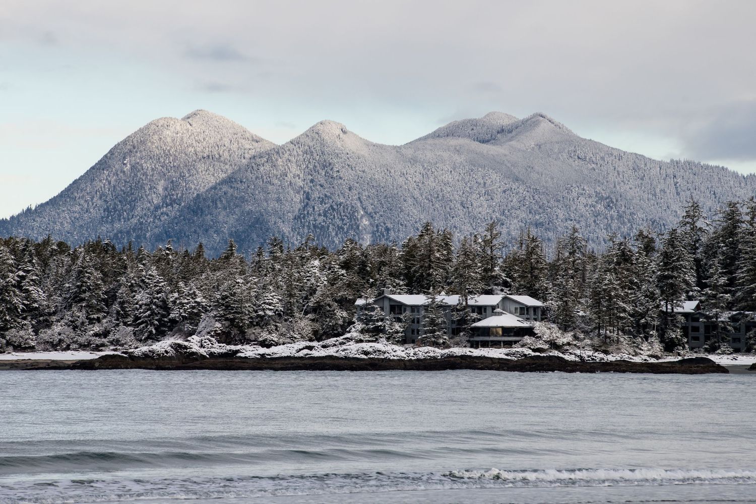 Snow-dusted mountains and evergreen forests line a peaceful winter harbor with waterfront structures.