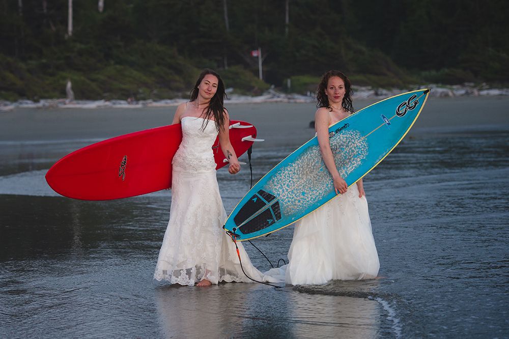 Two brides in white wedding dresses pose with surfboards on a beach at sunset.
