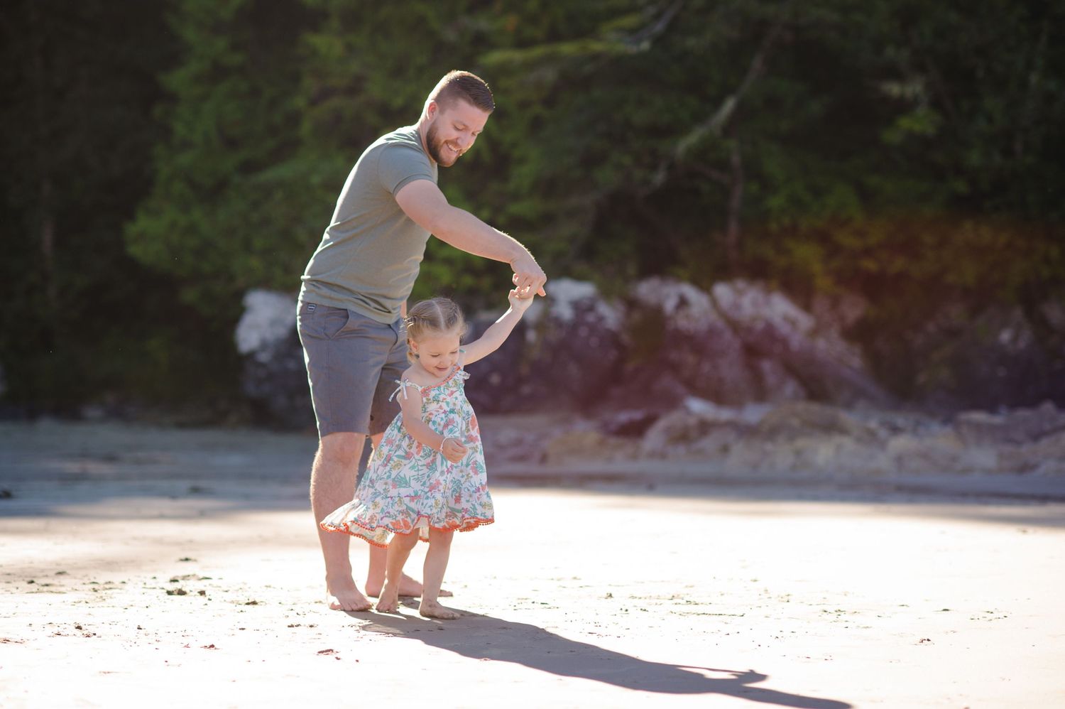 Young child in a floral dress twirls playfully on a sunny beach with rocky cliffs in the background.