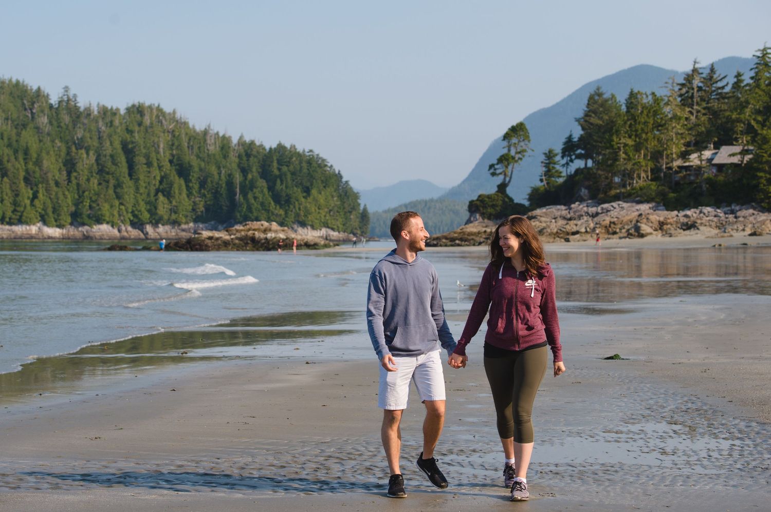Beachgoers stroll along a sandy Pacific Northwest coastline with forested mountains and clear blue water.
