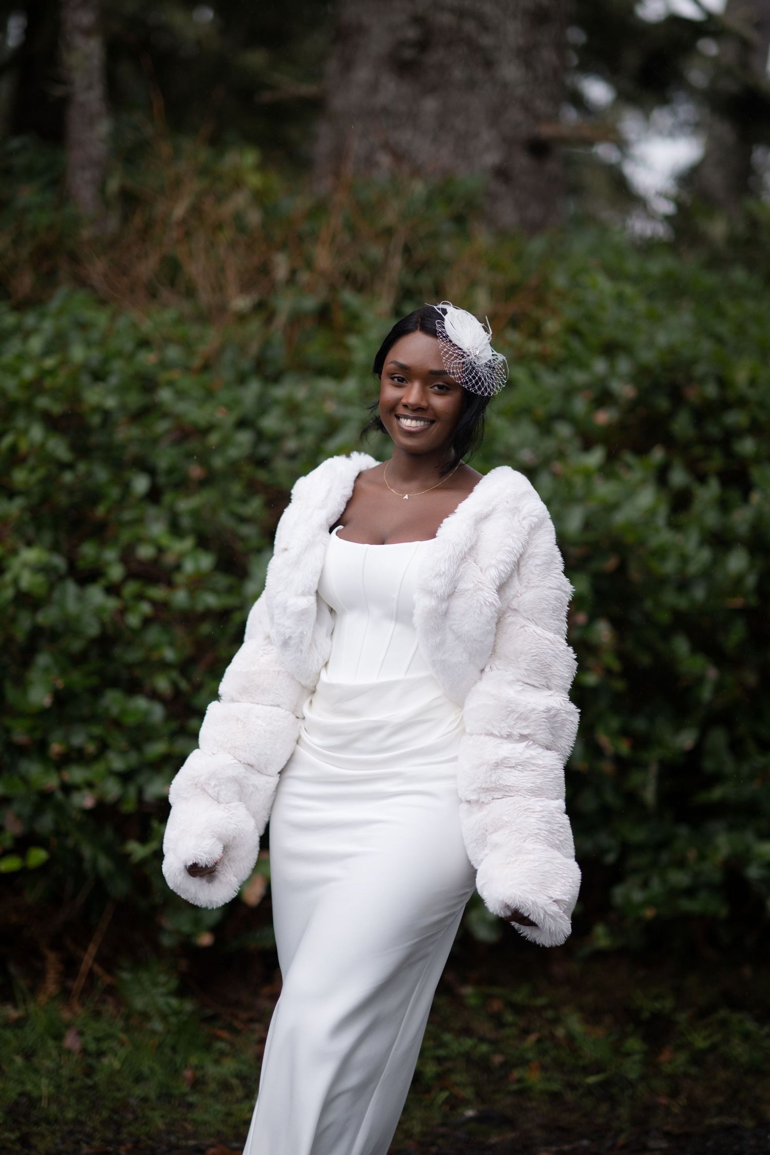A bride in an elegant white dress and fur sleeves poses in front of greenery during a winter photoshoot.