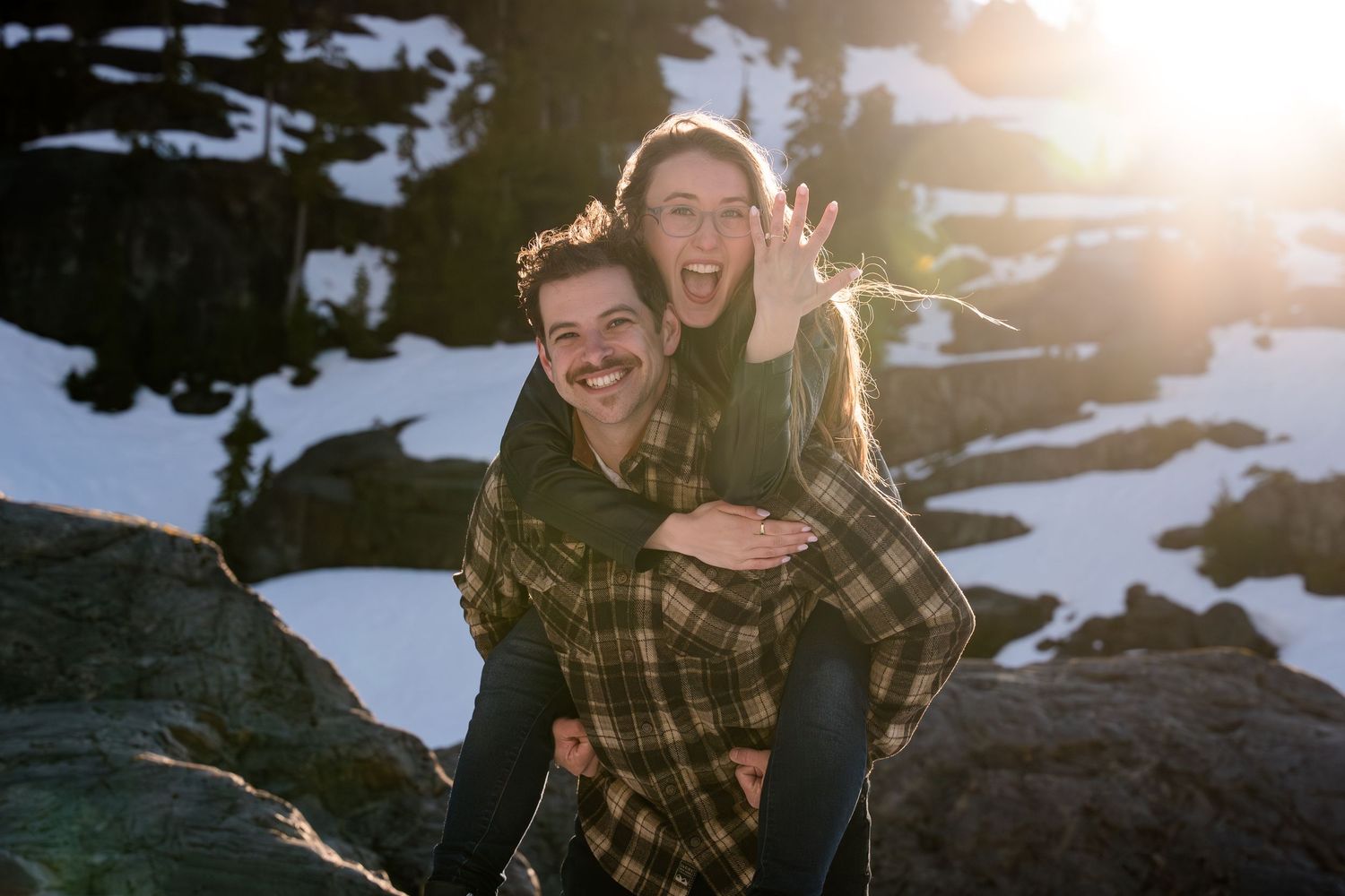 Couple laughing outdoors in snowy mountain setting, woman on man's back, both wearing plaid shirts, sun flare behind them.
