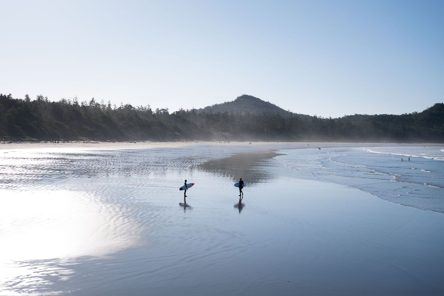 Distant surfers stand in calm waters of a scenic bay surrounded by forested hills on a tranquil morning.
