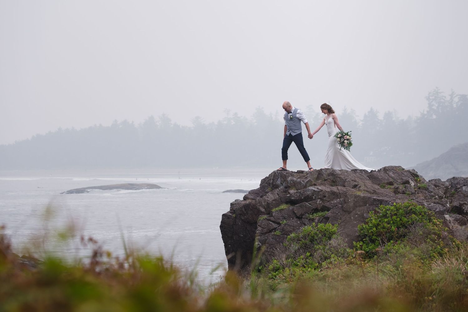 A wedding couple stands on a dramatic coastal cliff overlooking misty ocean views.