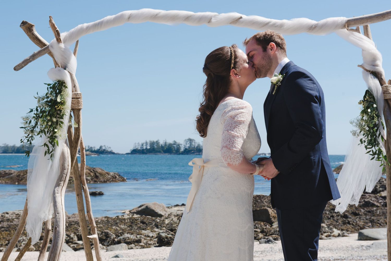 Rustic driftwood wedding arch decorated with white fabric stands on a rocky beach overlooking the ocean.