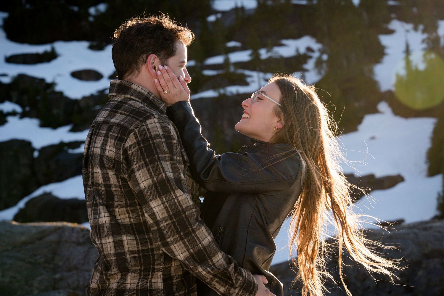 Joyful couple laughing and sharing a tender moment together on snowy mountain rocks at sunset.