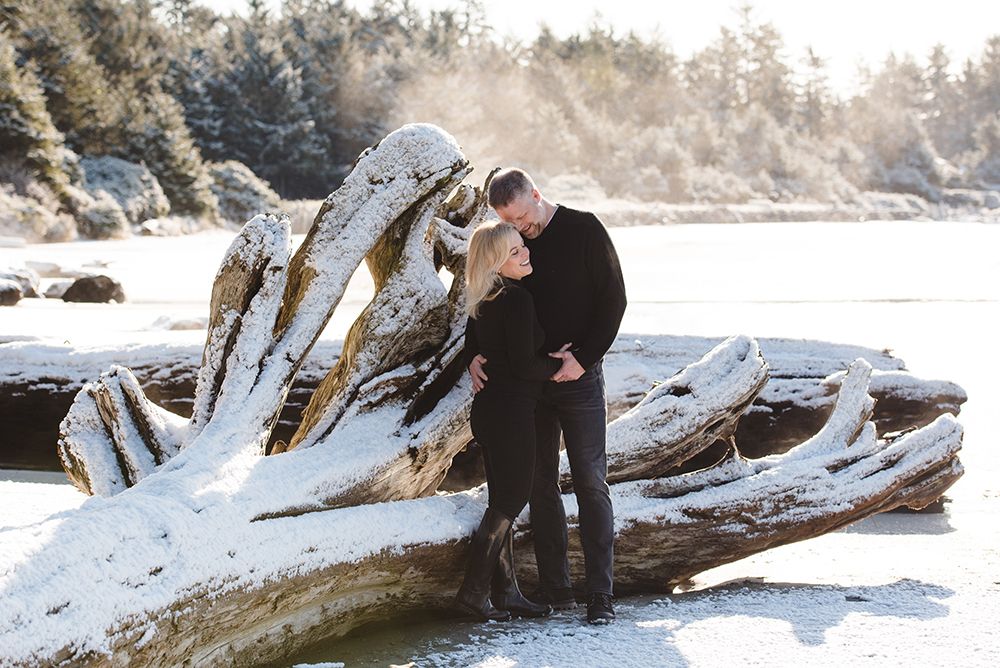 A couple embraces on a snow-covered driftwood log at the beach during winter.