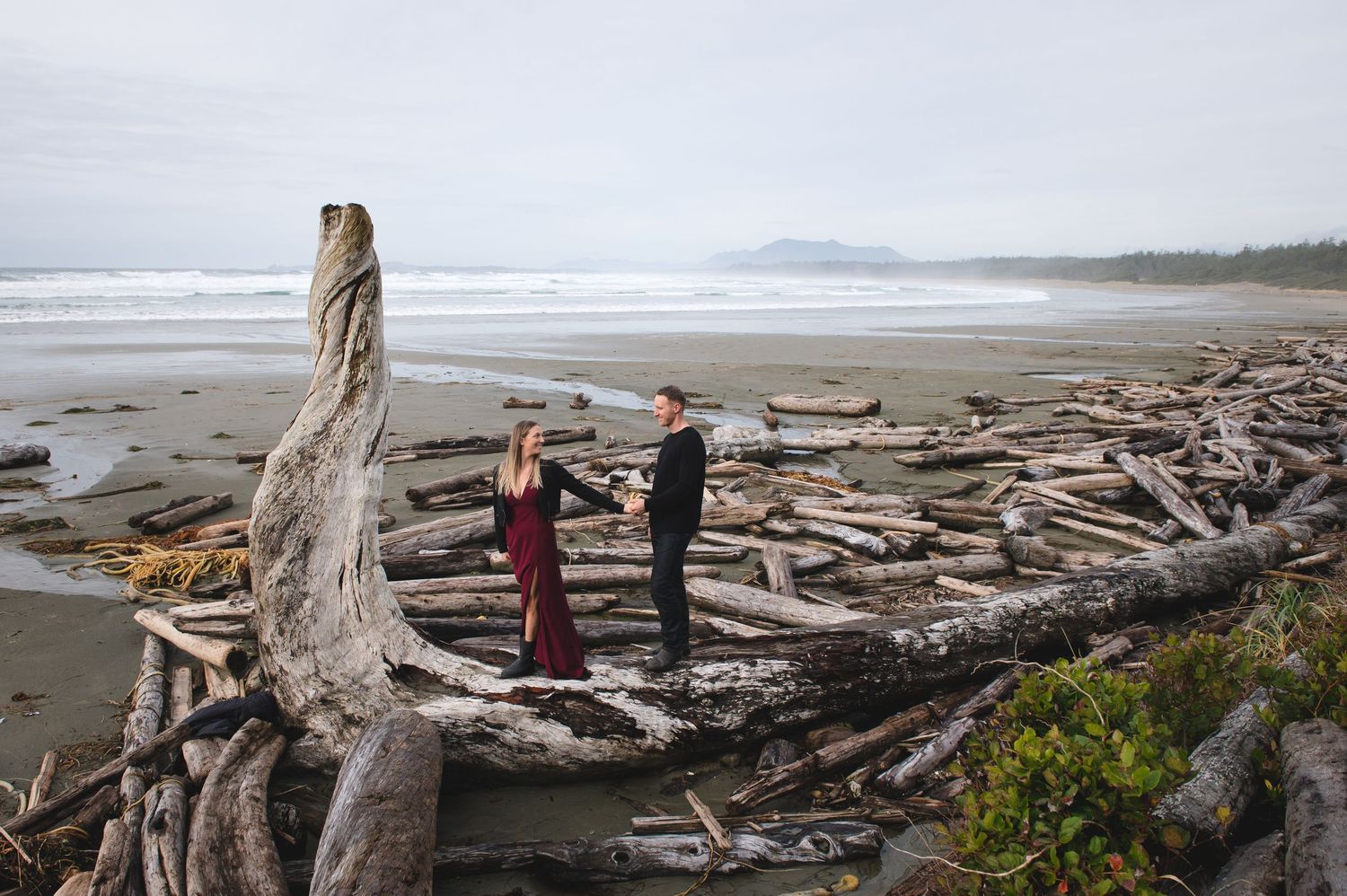 Two people explore a driftwood-covered beach with dramatic coastal scenery.