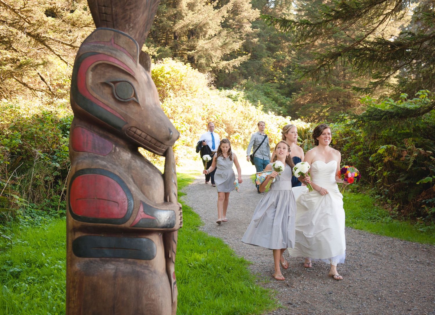 Traditional totem pole stands along a forest path where people walk in white attire.