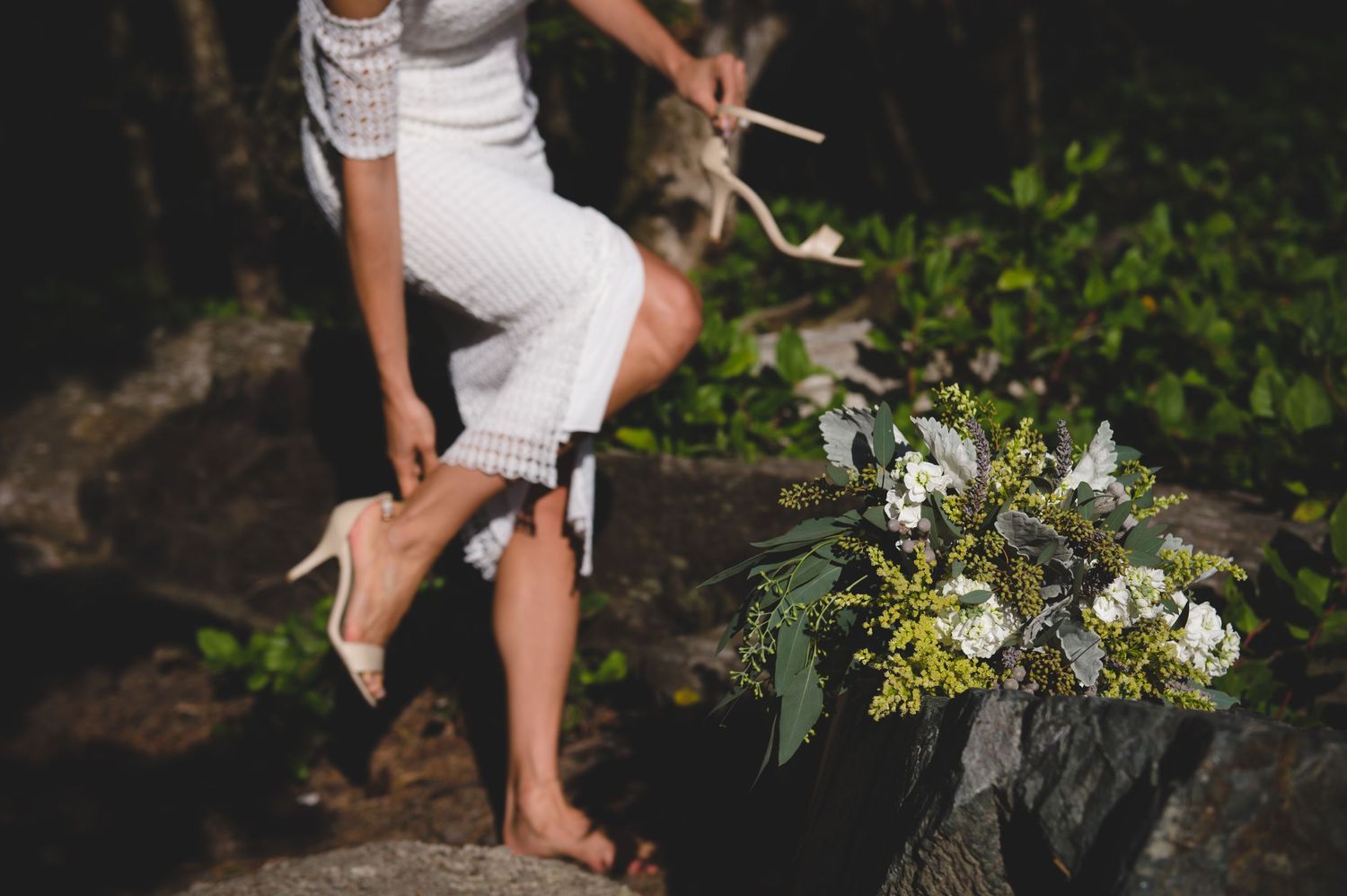 People walking barefoot on rocky beach surrounded by lush greenery and white flowers.
