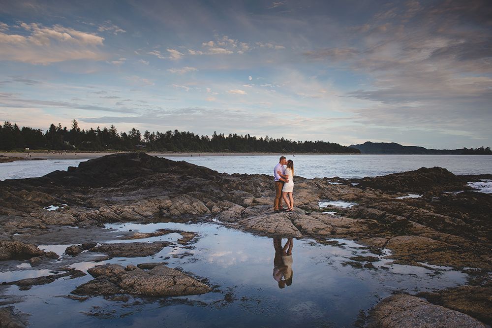 A person stands on rocky coastal shores during sunset with dramatic cloudy skies reflecting in tidal pools.