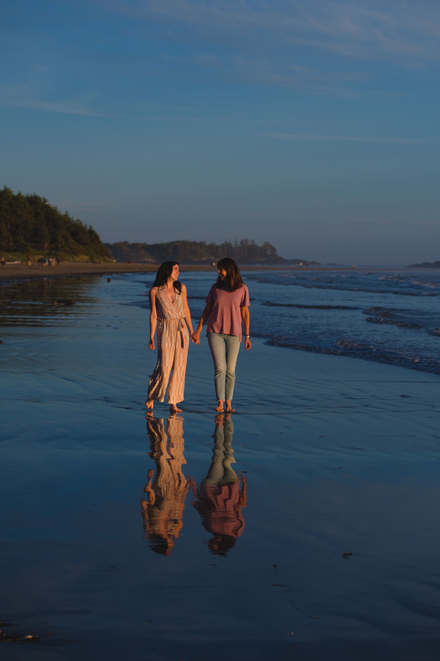 Two people walking along a scenic beach at sunset with their reflections in the wet sand.