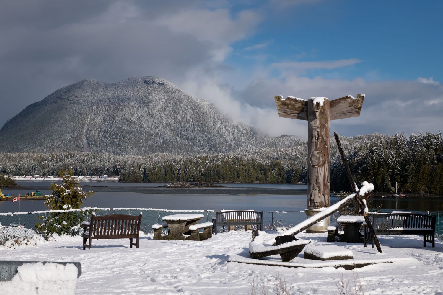 Snow-covered benches and trees line a scenic mountain lake view under cloudy winter skies.