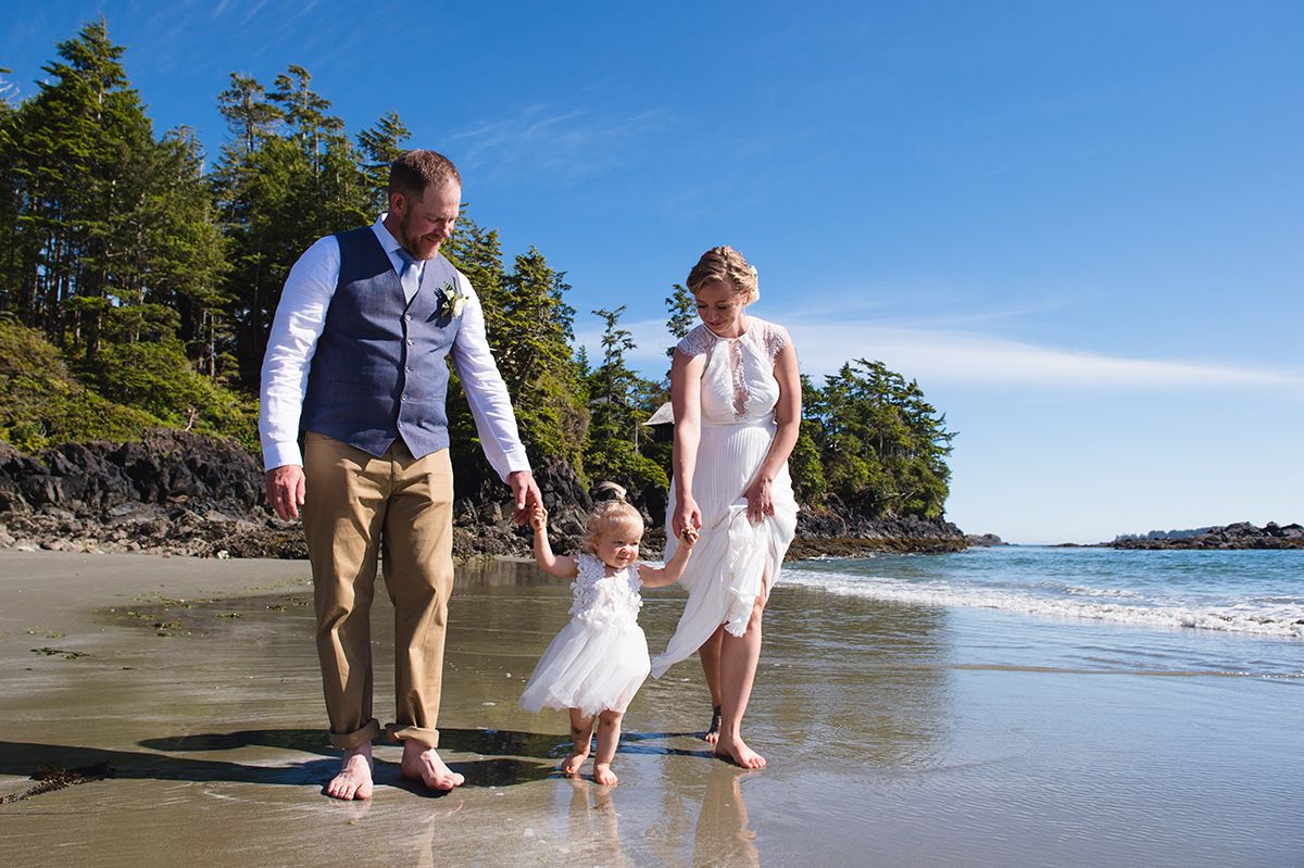 Wedding party walking along a scenic beach shoreline with evergreen trees in the background.