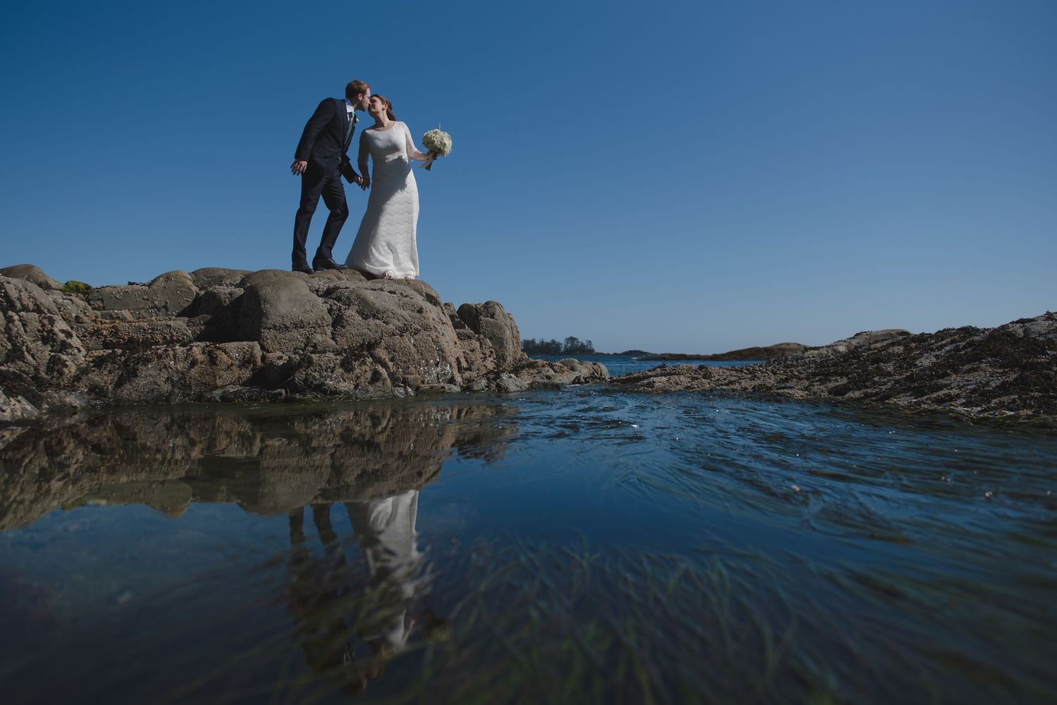 Wedding couple reflected in tidal pools on rocky beach with blue sky.