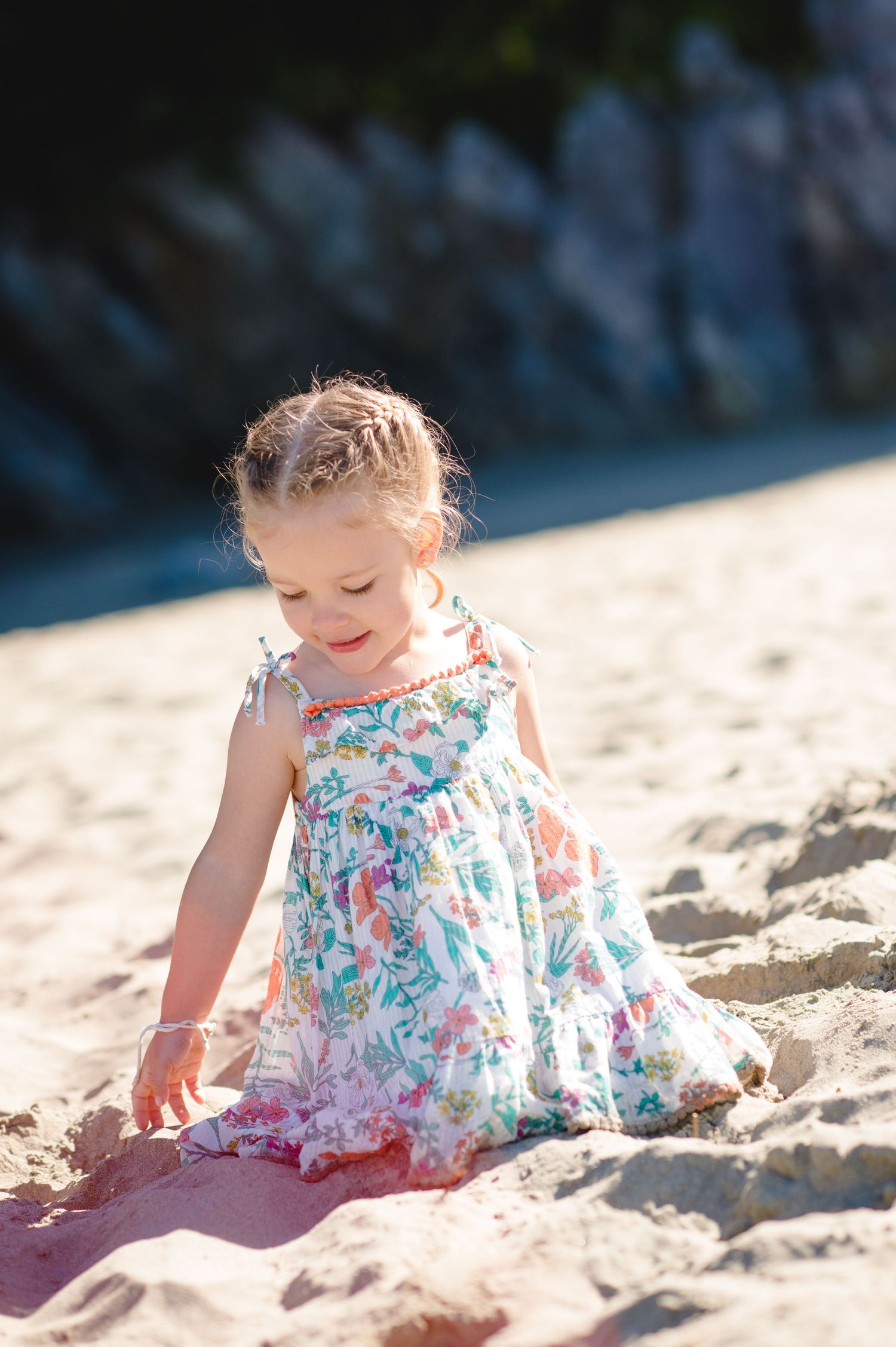 Child in floral dress playing joyfully in beach sand during summer afternoon.
