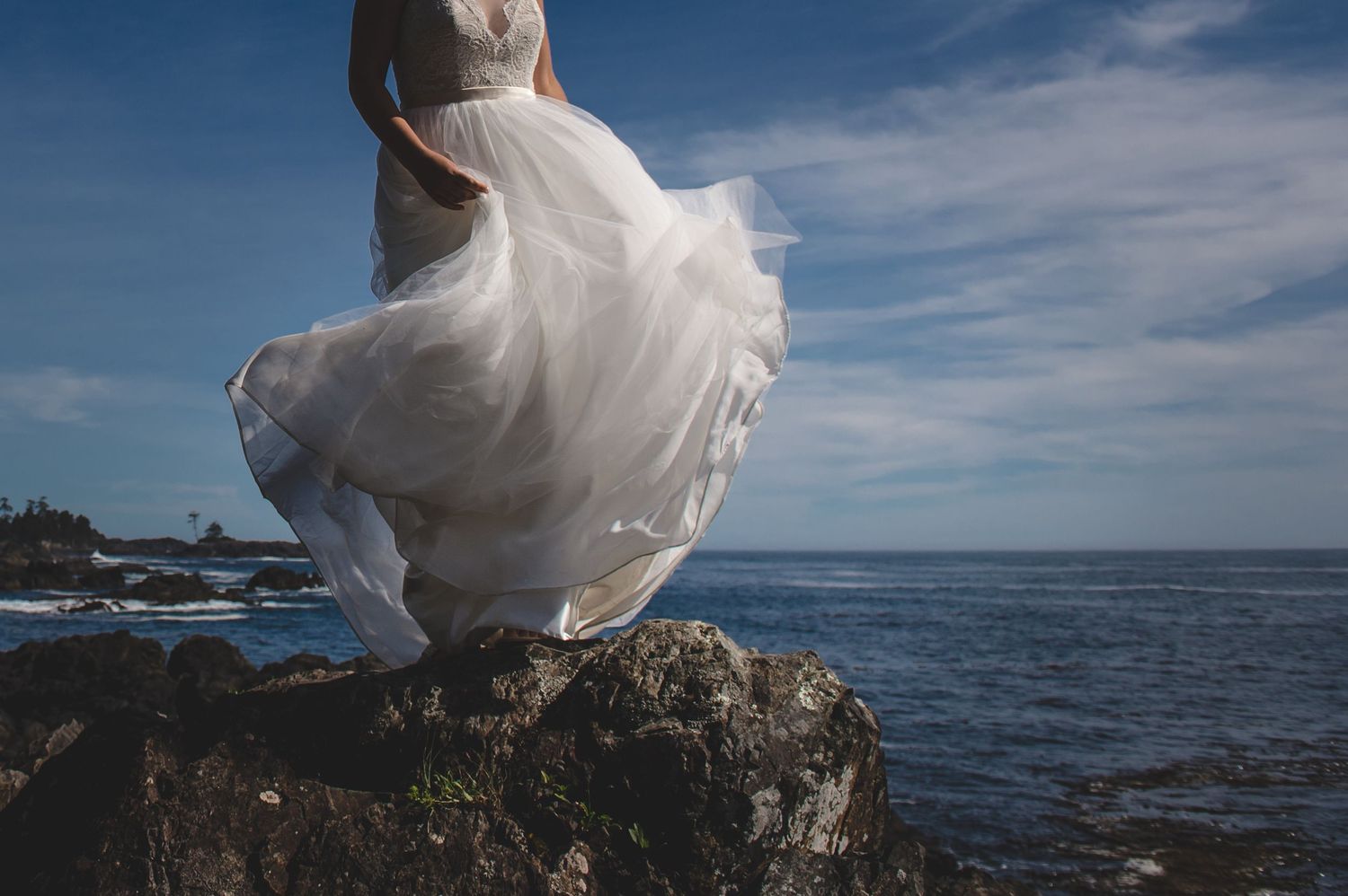 White wedding dress flows in the wind against dramatic ocean and rocky coastline backdrop.