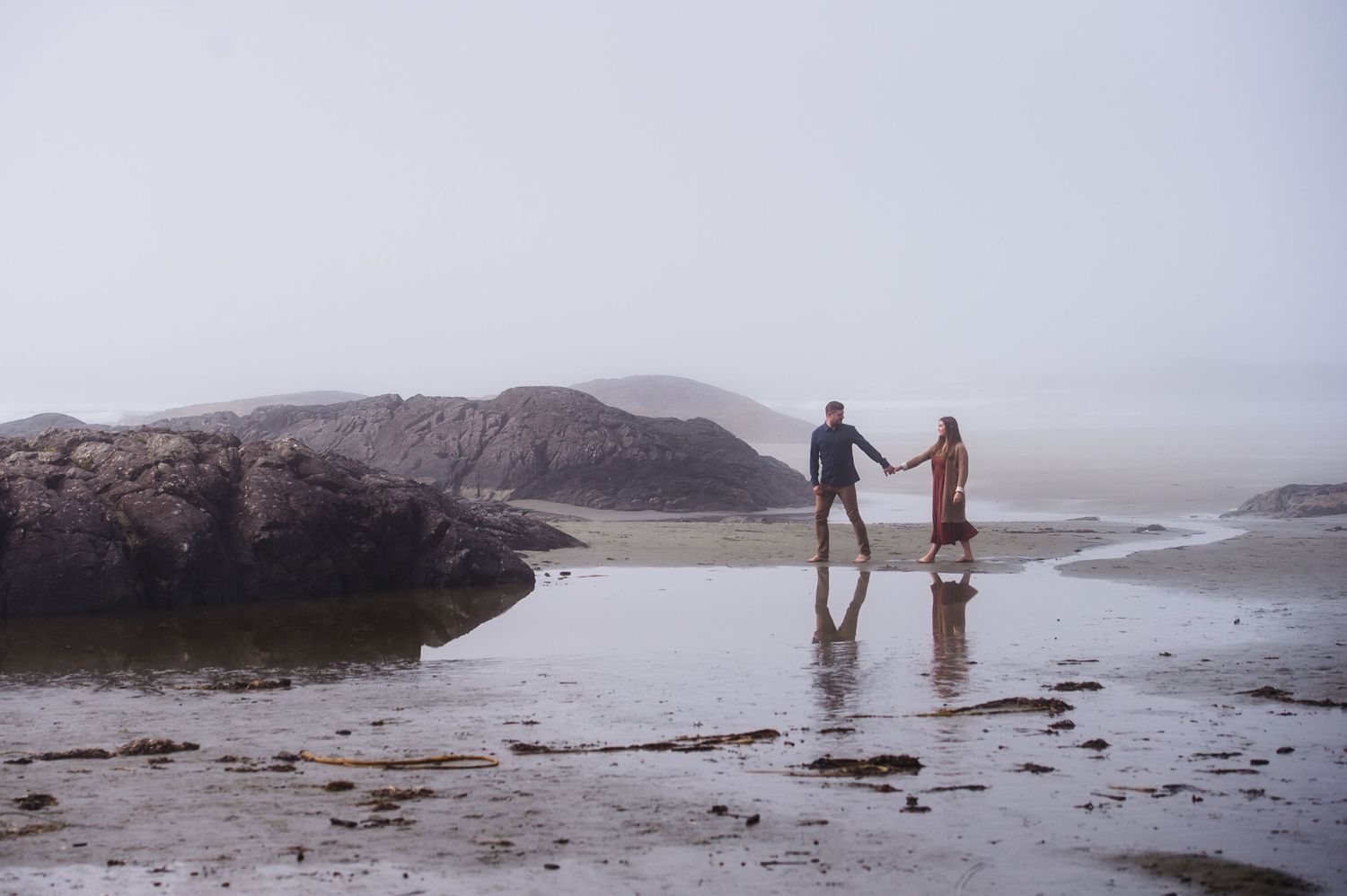 People exploring rocky coastline during overcast day with mountains visible in distance.