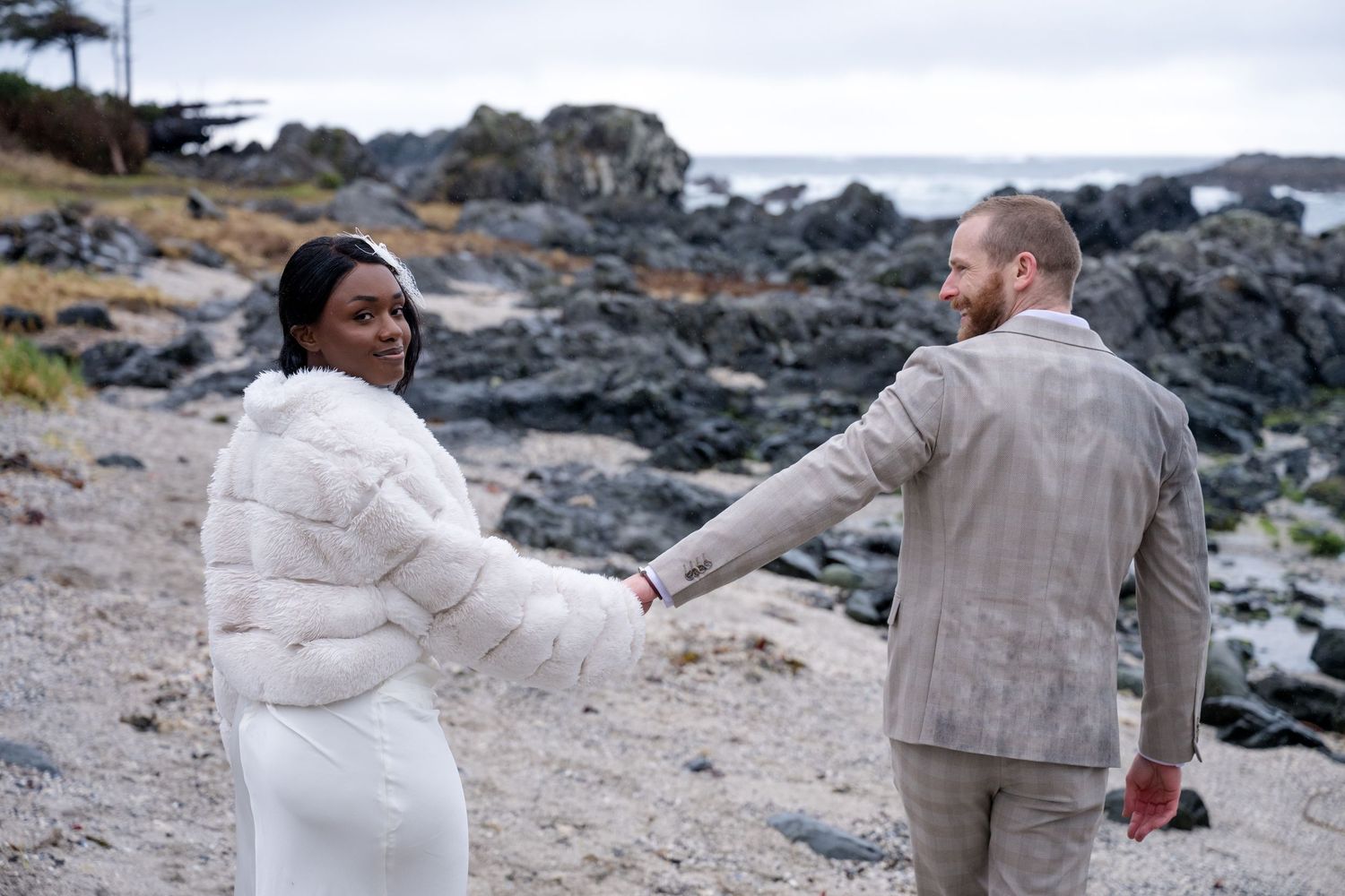 A couple sits together on rocky coastal terrain with ocean waves and rugged shoreline in the background.