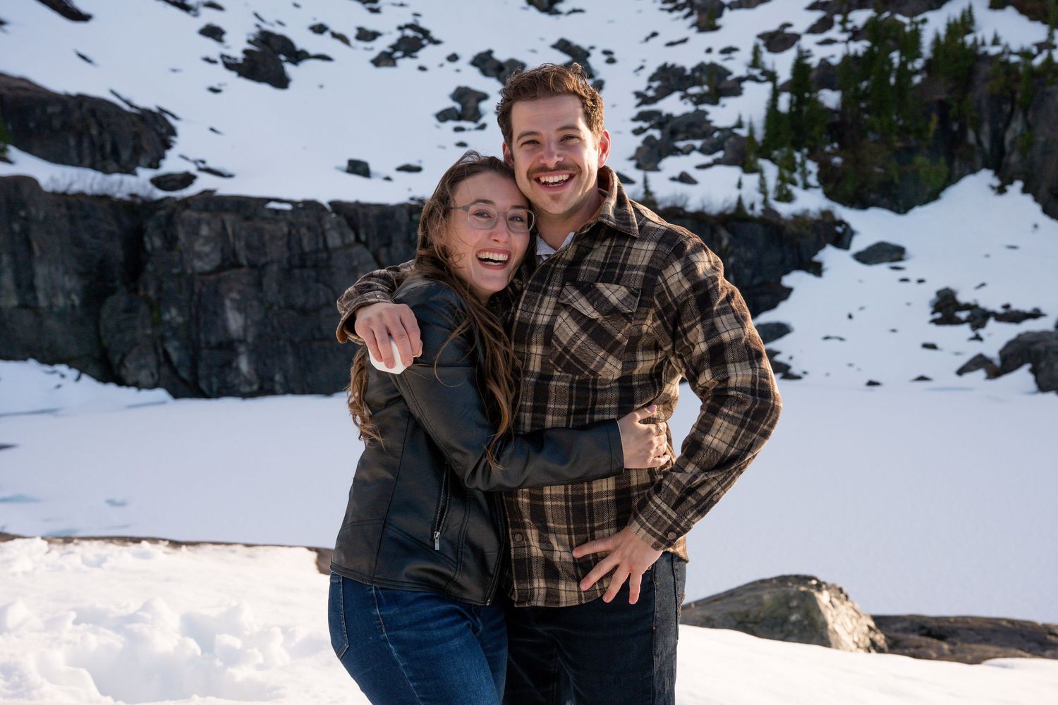 Happy couple embracing and smiling together against a stunning snowy mountain backdrop during engagement photos.