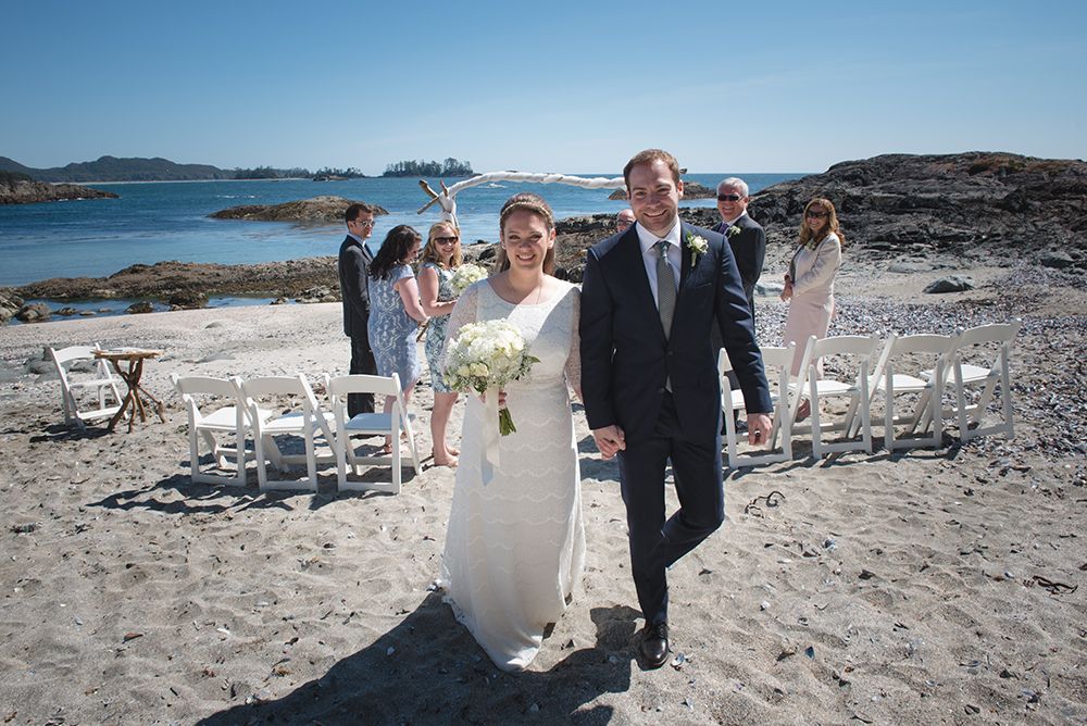Wedding guests seated on white chairs at a rocky beach ceremony site with ocean views.