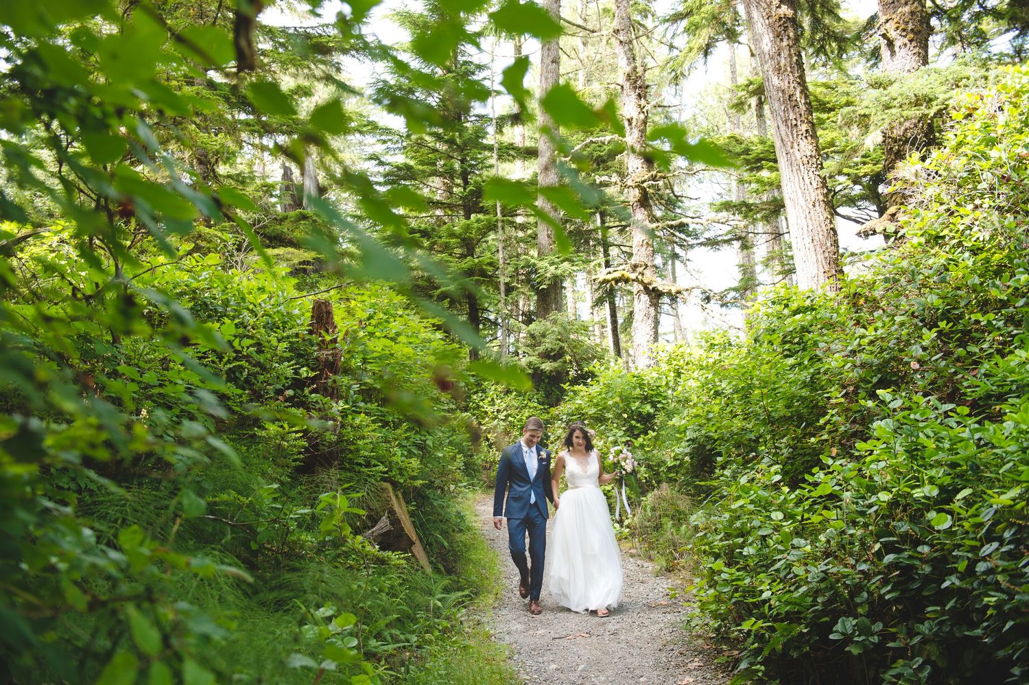A couple walks down a lush forest path surrounded by green foliage.