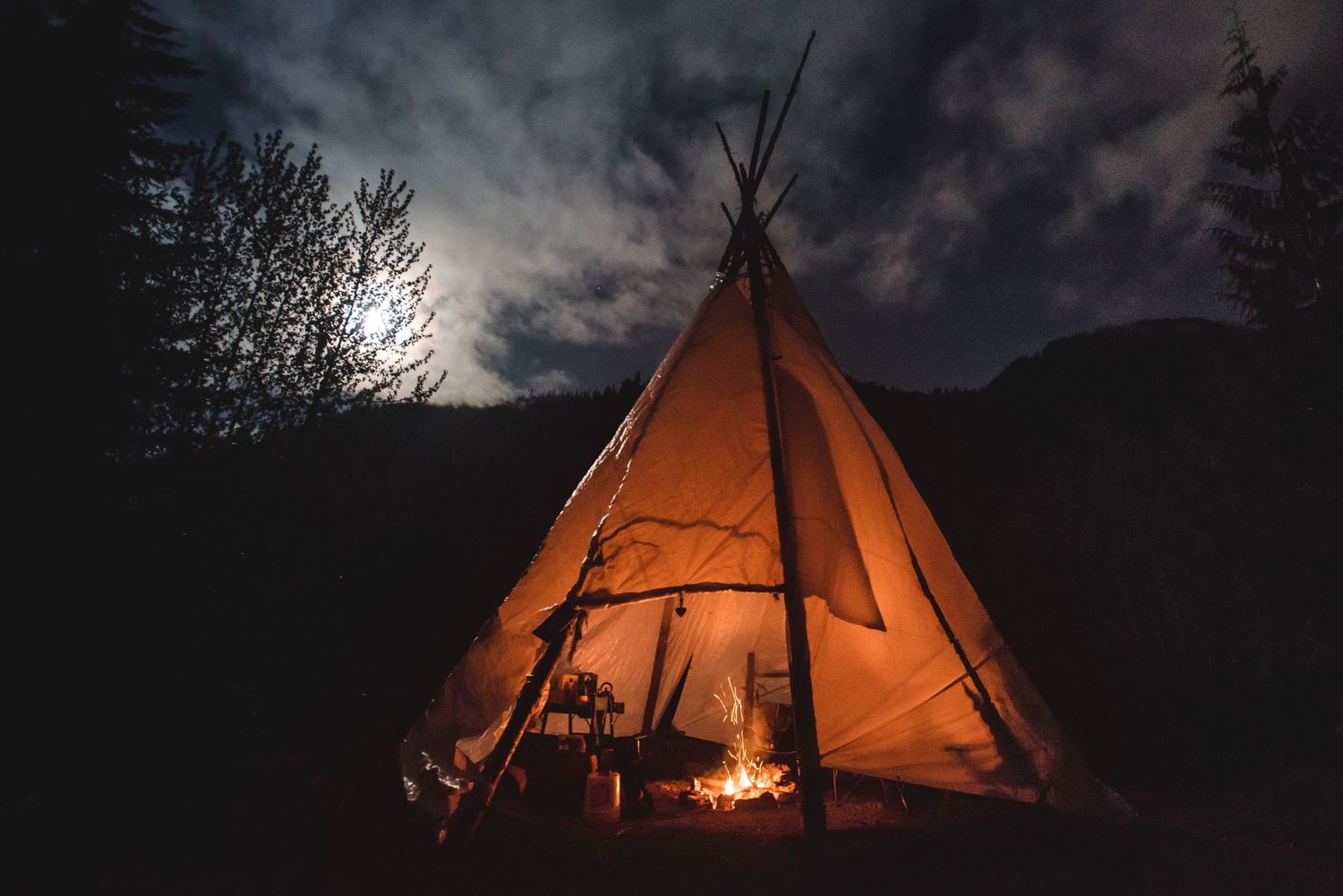 Fairy Creek blockade. Illuminated teepees glowing orange against dark night sky with moonlight breaking through clouds.