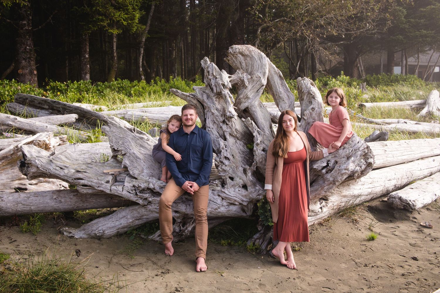 A family poses together on a large piece of driftwood at the beach during golden hour.