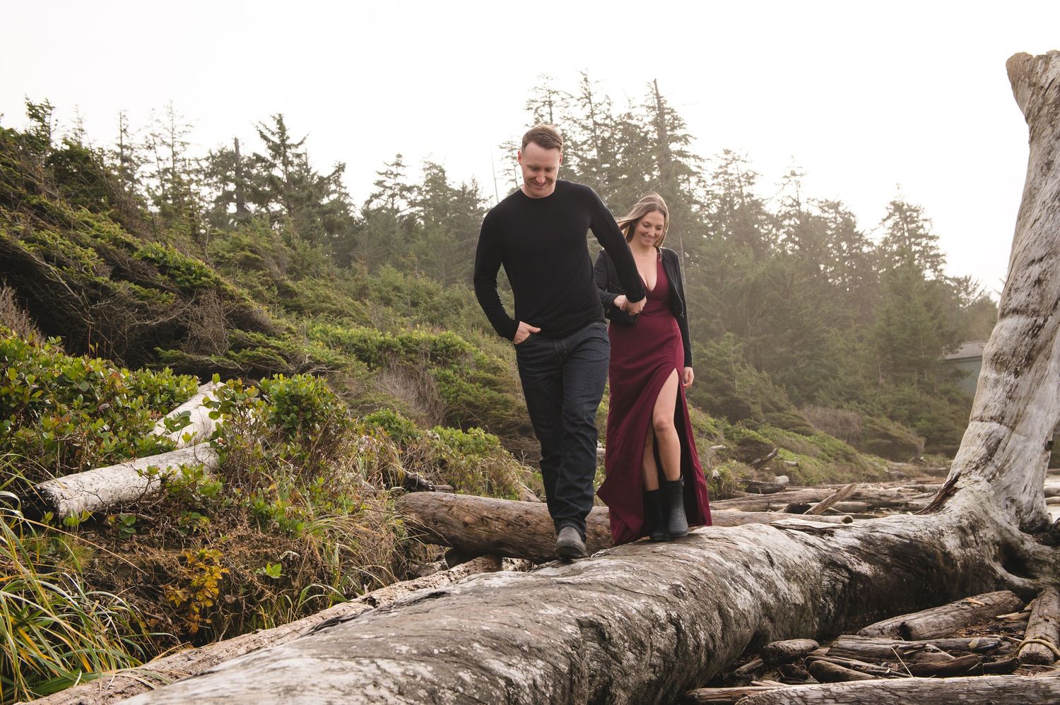 Couple walking among weathered driftwood on rugged Pacific coast beach.
