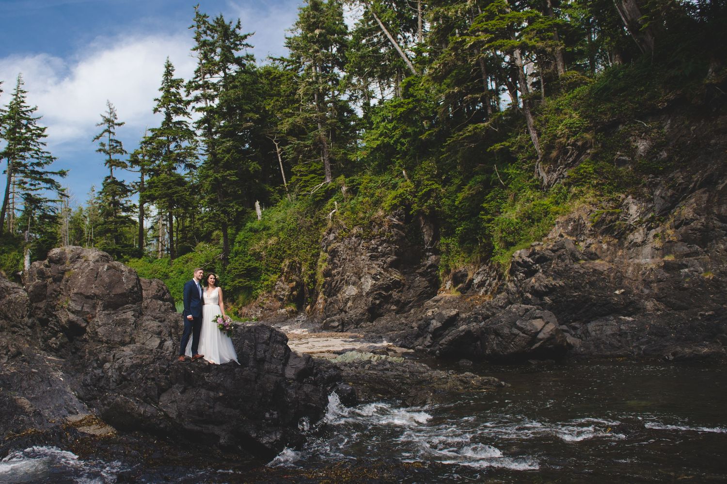 A couple stands on rugged coastal rocks surrounded by evergreen trees and crashing waves along the Pacific Northwest shore.