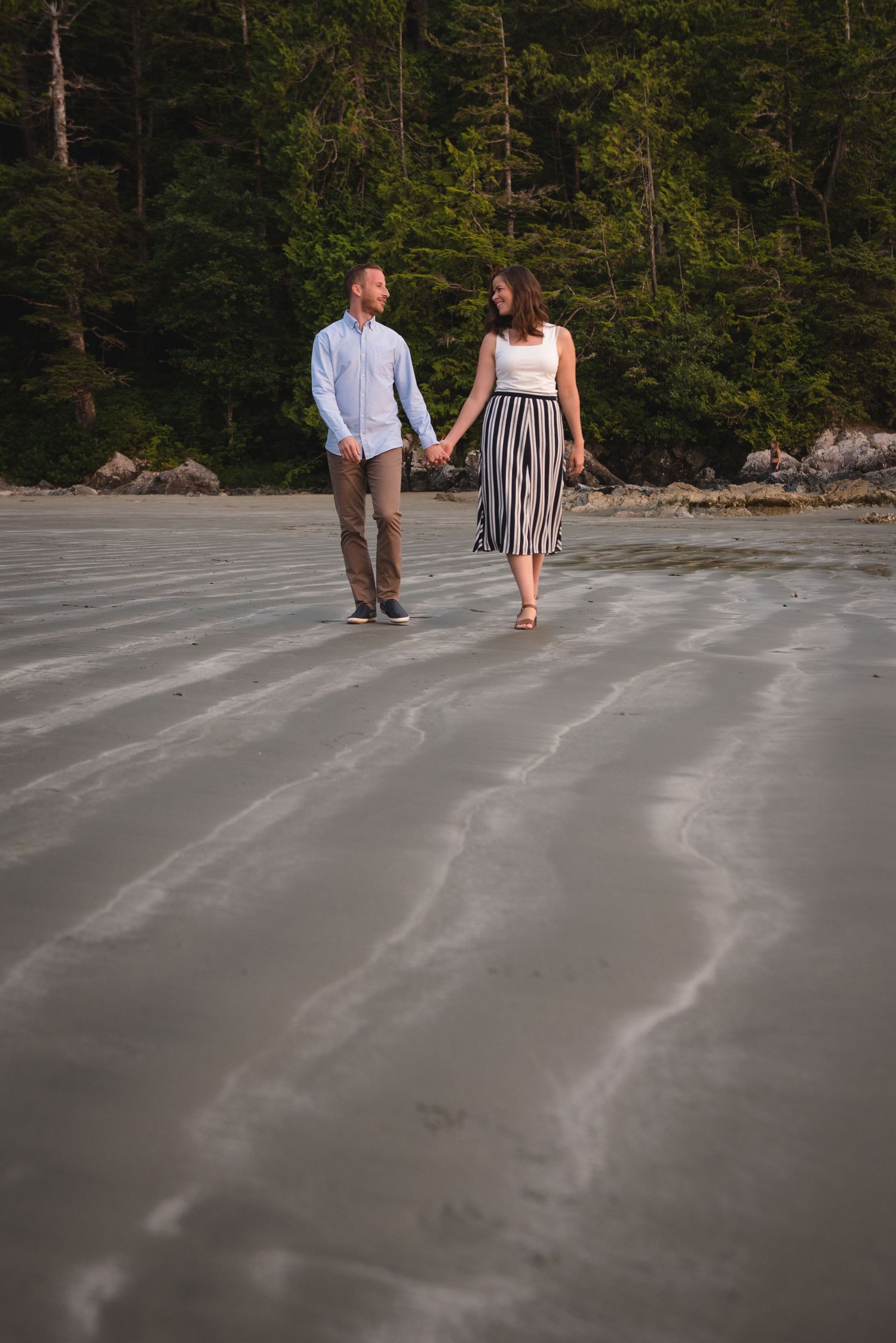 Couple walks hand in hand on concrete surface with forest backdrop at sunset.