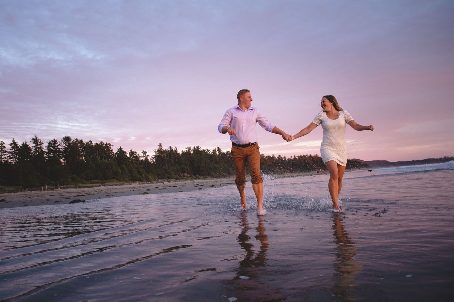 Two people holding hands while walking along a beach shoreline at sunset with reflections in the wet sand.