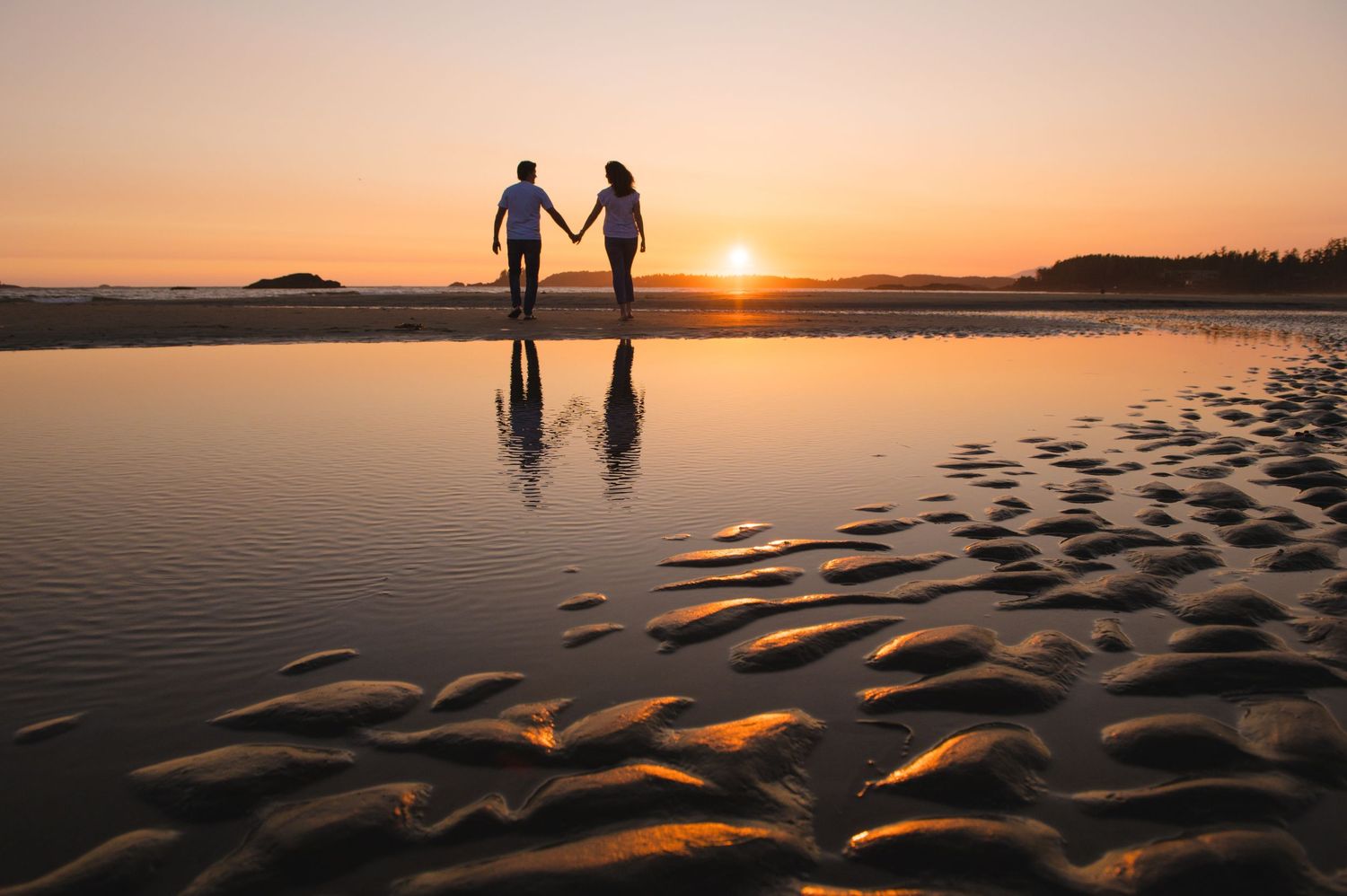Two silhouettes hold hands on a beach at sunset with rippled sand and mountains in the background.