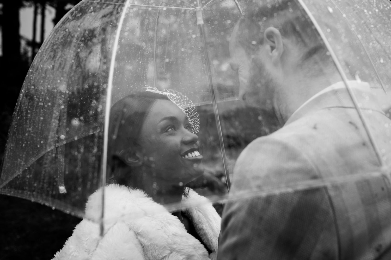 A couple embraces beneath clear umbrellas while rain falls around them during an outdoor photoshoot.