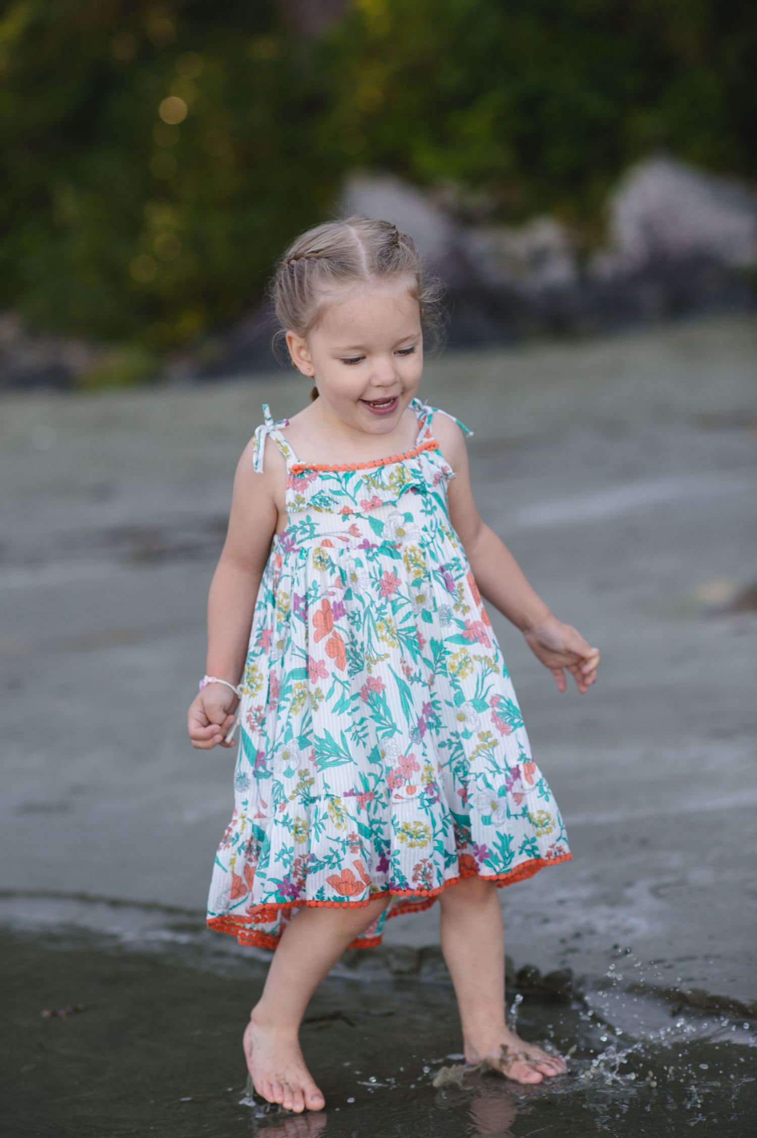 A small child in a floral dress splashes and plays at the water's edge on a sunny day.