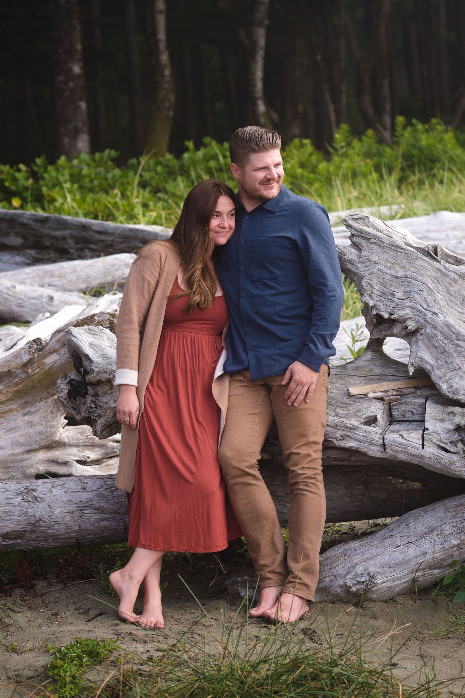 A couple stands barefoot together on driftwood at the beach near a forest.