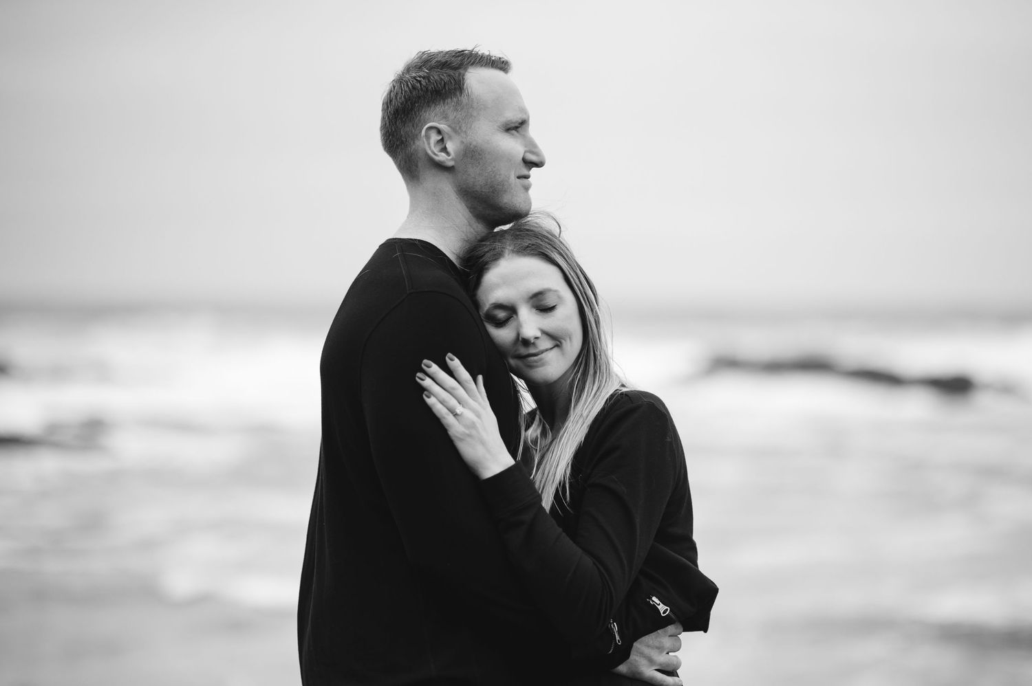 Intimate black and white portrait of couple embracing on cloudy beach.