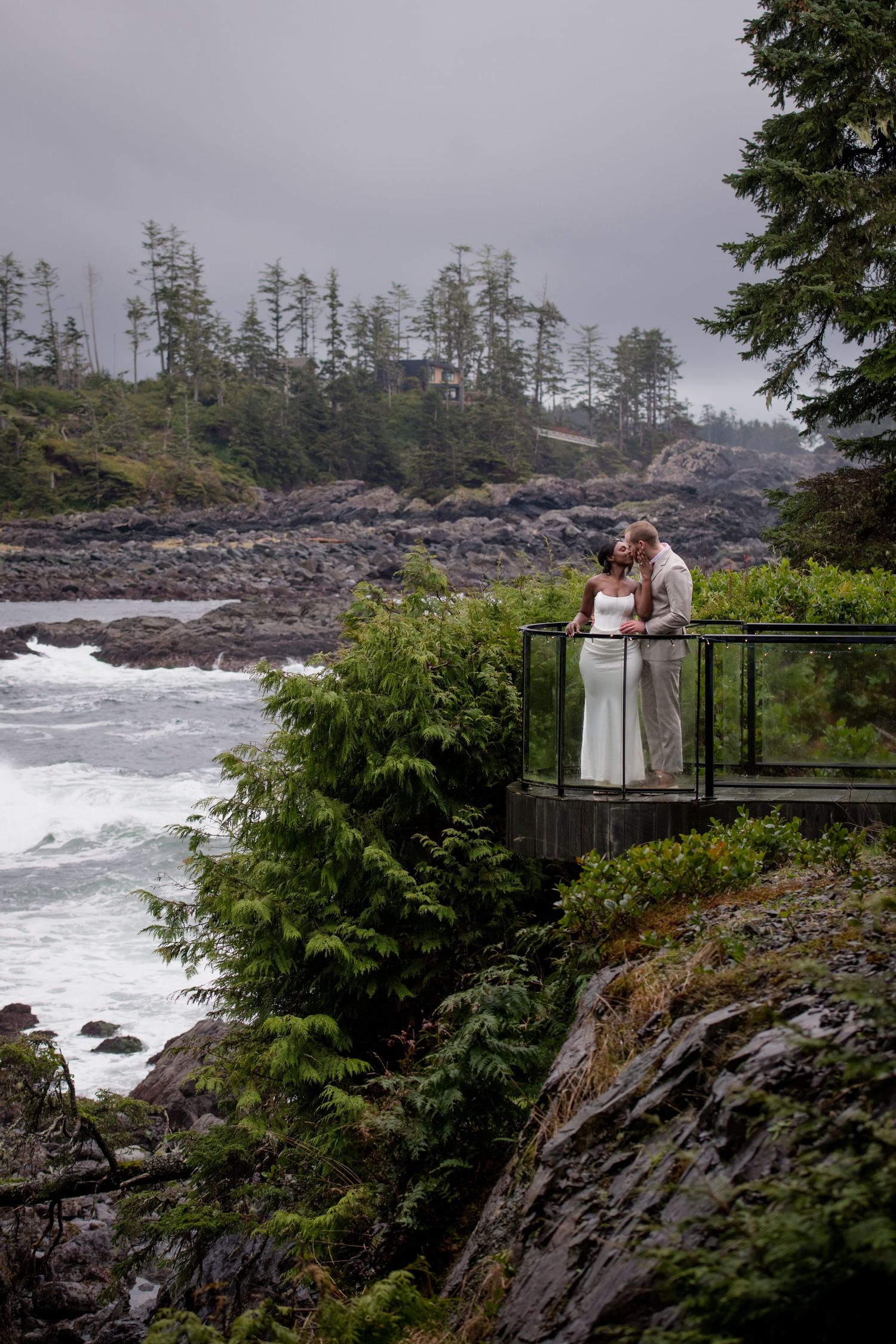 Dramatic coastal wedding photography on rugged cliffs overlooking the Pacific Ocean with crashing waves and evergreen trees.