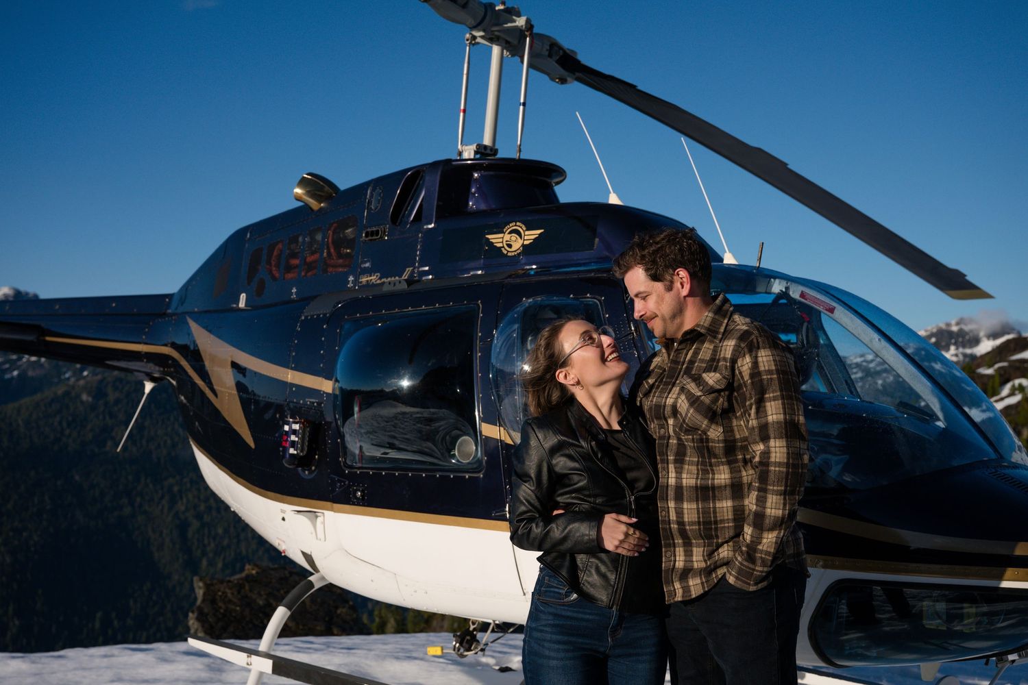 Romantic couple embracing in front of an Atleo Air helicopter on a snow-covered mountain summit in bright sunlight.