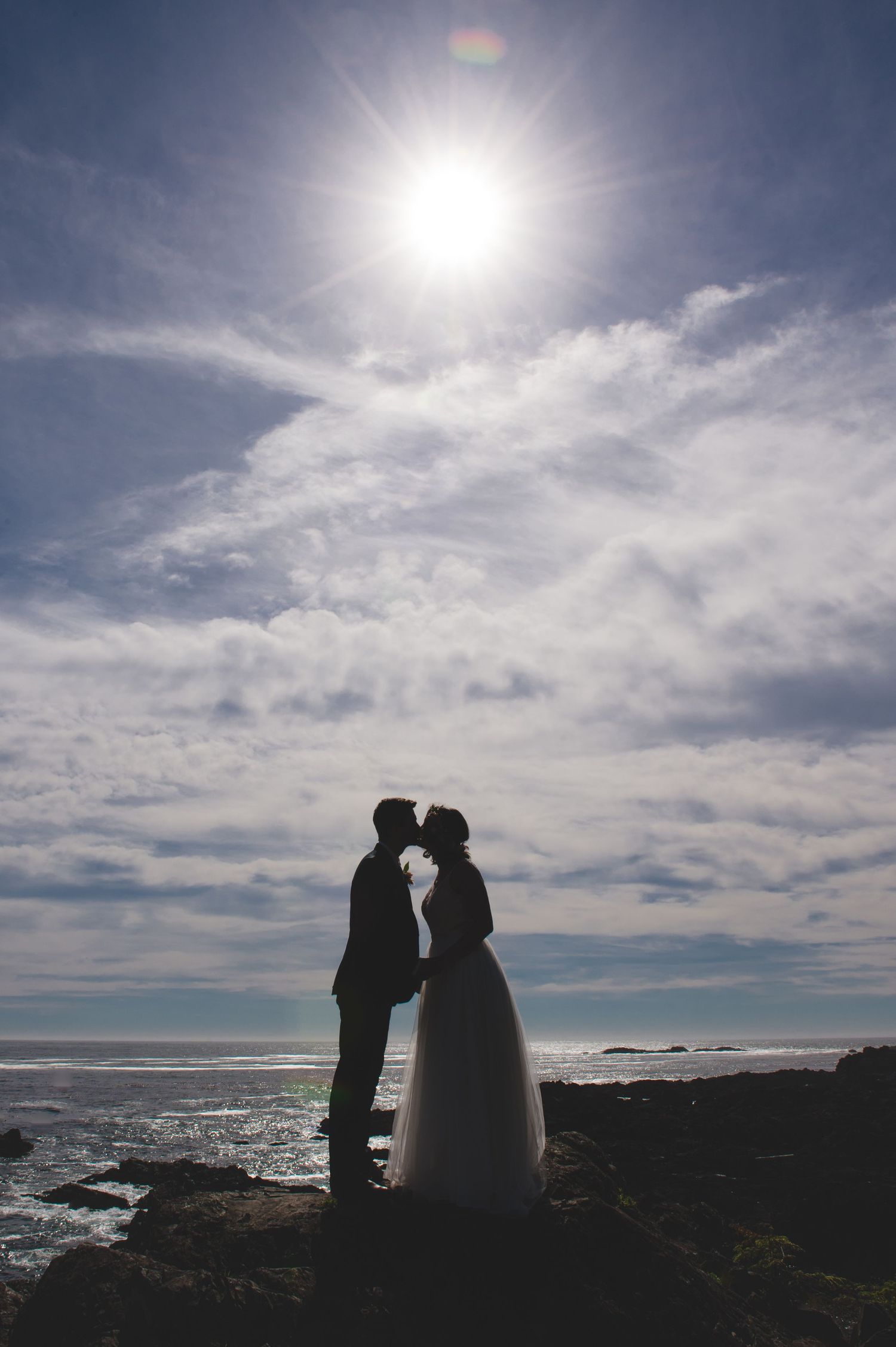Silhouetted wedding couple against bright sun and ocean horizon on rocky shore.