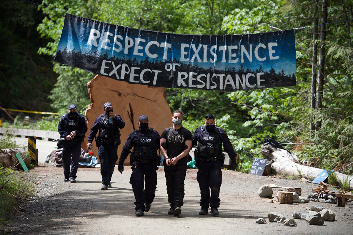 Fairy Creek blockade. Police officers walk under a blue banner reading 'Respect Existence Expect Resistance' on a forest path.